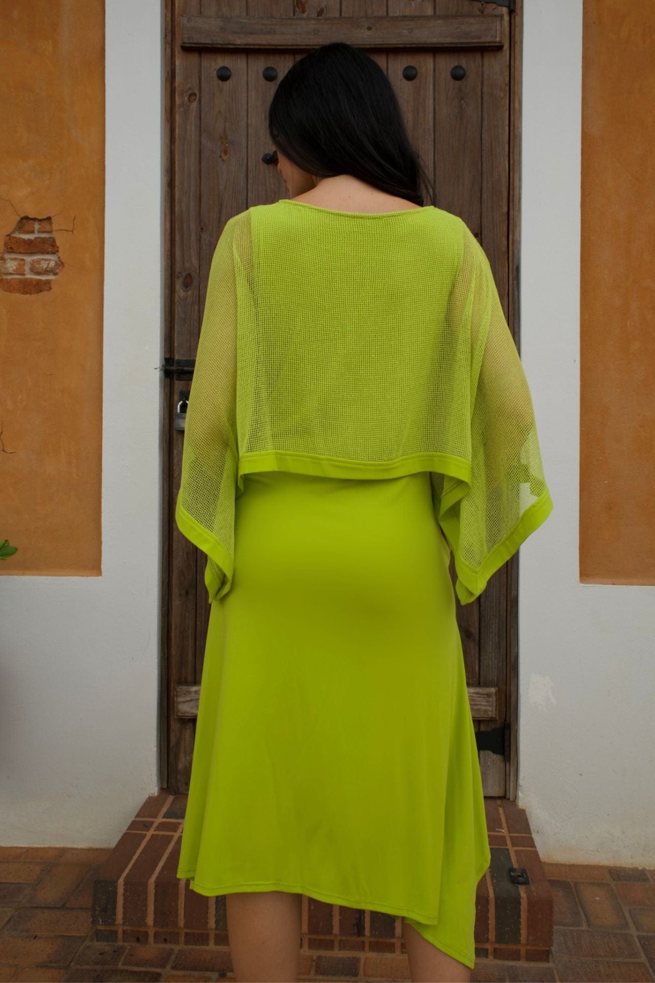 Woman wearing a lime green sleeveless jersey dress with a mesh overlay, standing in front of a rustic wooden door.