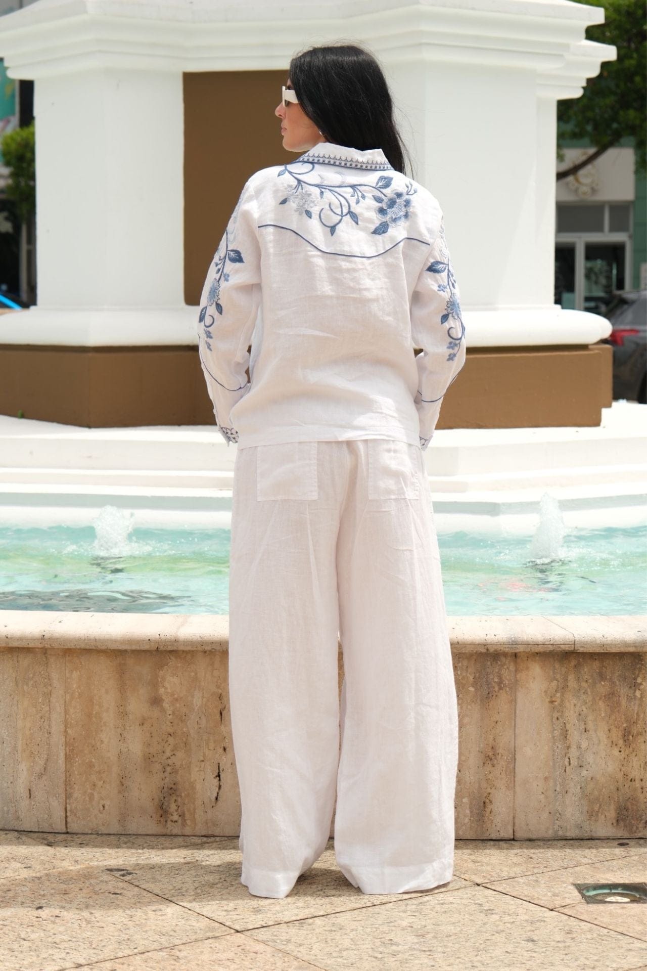 Back view of a model wearing white and blue embroidered linen set with relaxed fit top and wide-leg pants.