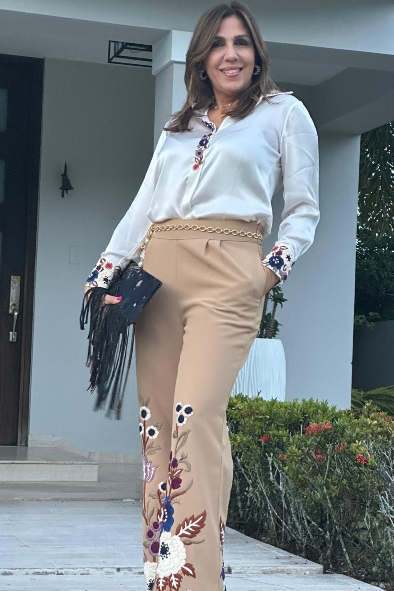 Woman wearing beige embroidered pant set with white blouse, styled with a gold chain belt and black fringe clutch — an elegant, elevated look completed with sophisticated accessories.

