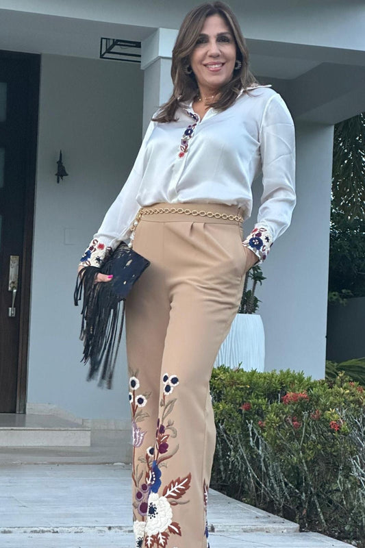 Woman wearing beige embroidered pant set with white blouse, styled with a gold chain belt and black fringe clutch — an elegant, elevated look completed with sophisticated accessories.

