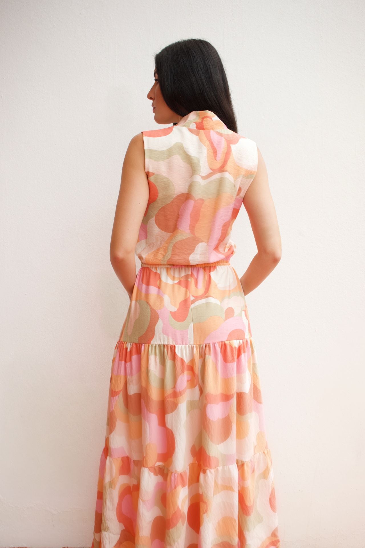 Woman wearing a sleeveless abstract print tiered maxi dress in soft coral and peach tones with an elastic waist and flowing silhouette.