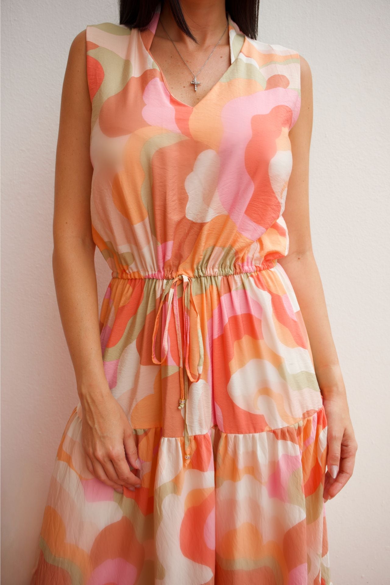Woman wearing a sleeveless abstract print tiered maxi dress in soft coral and peach tones with an elastic waist and flowing silhouette.