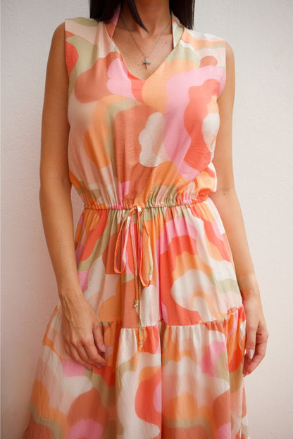 Woman wearing a sleeveless abstract print tiered maxi dress in soft coral and peach tones with an elastic waist and flowing silhouette.