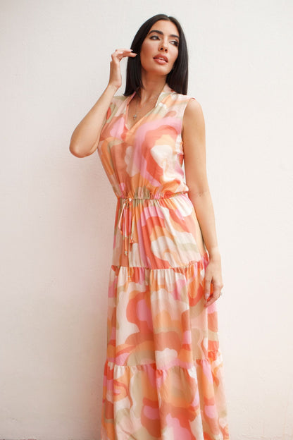 Woman wearing a sleeveless abstract print tiered maxi dress in soft coral and peach tones with an elastic waist and flowing silhouette.