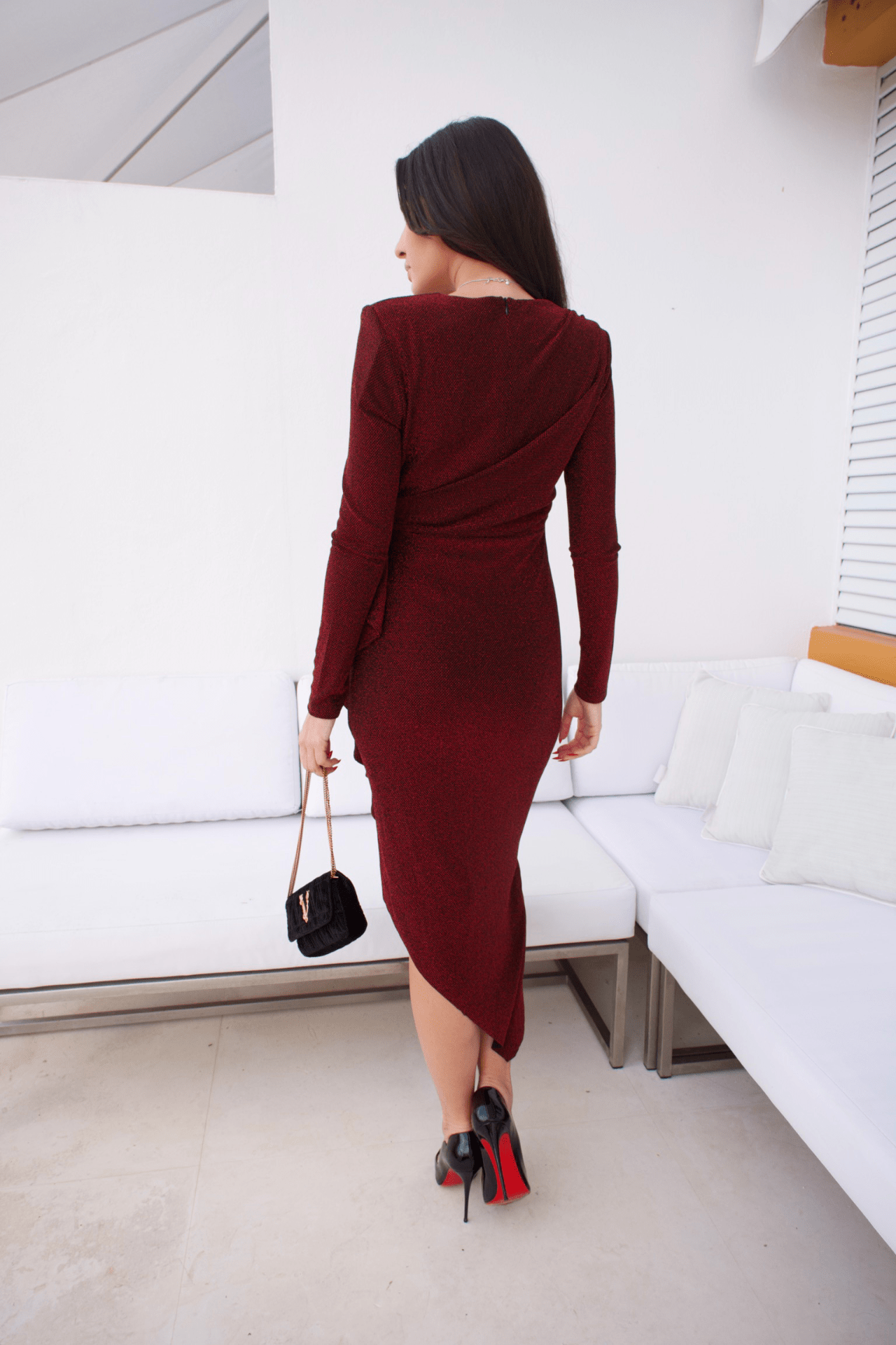 A woman wearing a red, asymmetrically designed metallic midi dress with a round neck and long ruched sleeves, standing in a room with white furniture and a handbag on her left side.