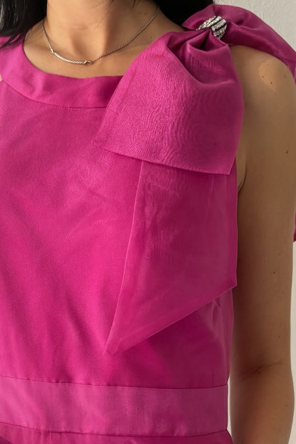 Woman wearing the Aurora Statement Jumpsuit in fuchsia with shoulder bow, wide-leg silhouette and sheer flowing overlay.