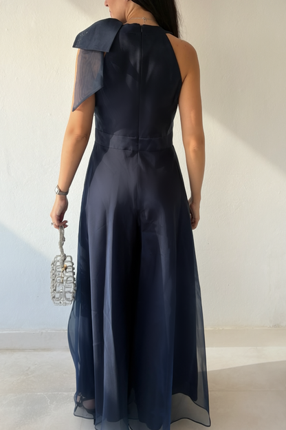 Woman wearing the Aurora Statement Jumpsuit in navy blue with shoulder bow, wide-leg silhouette and sheer flowing overlay.
