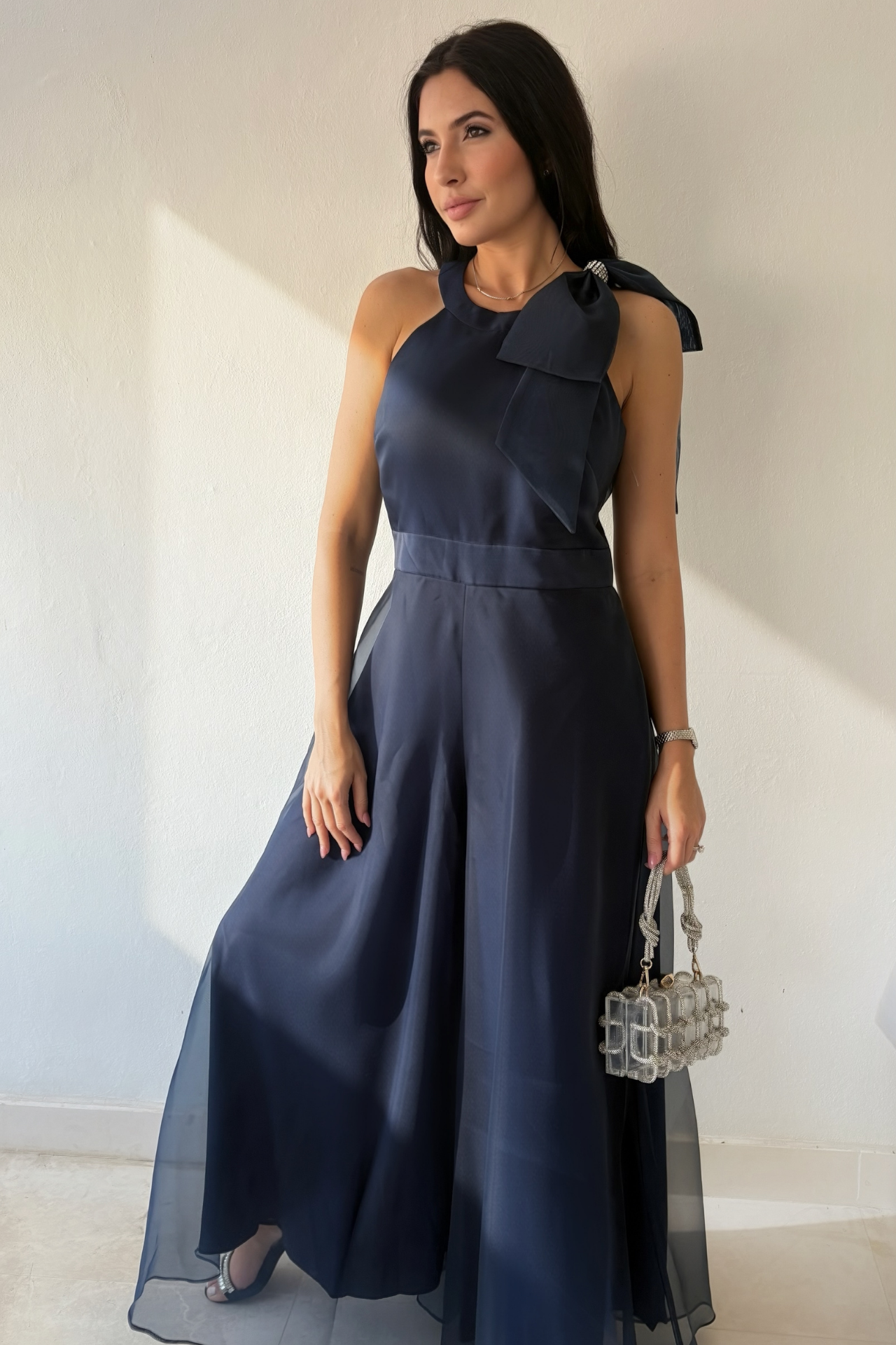 Woman wearing the Aurora Statement Jumpsuit in navy blue with shoulder bow, wide-leg silhouette and sheer flowing overlay.