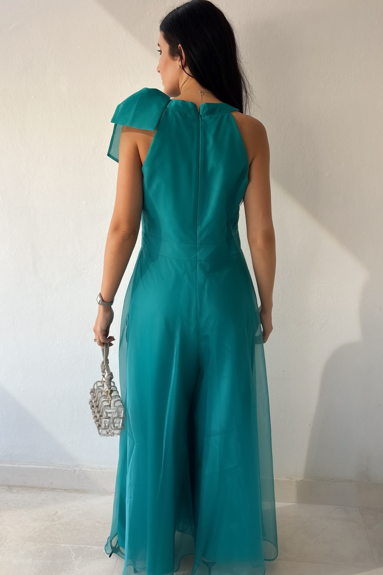Woman wearing the Aurora Statement Jumpsuit in teal with shoulder bow, wide-leg silhouette and sheer flowing overlay.