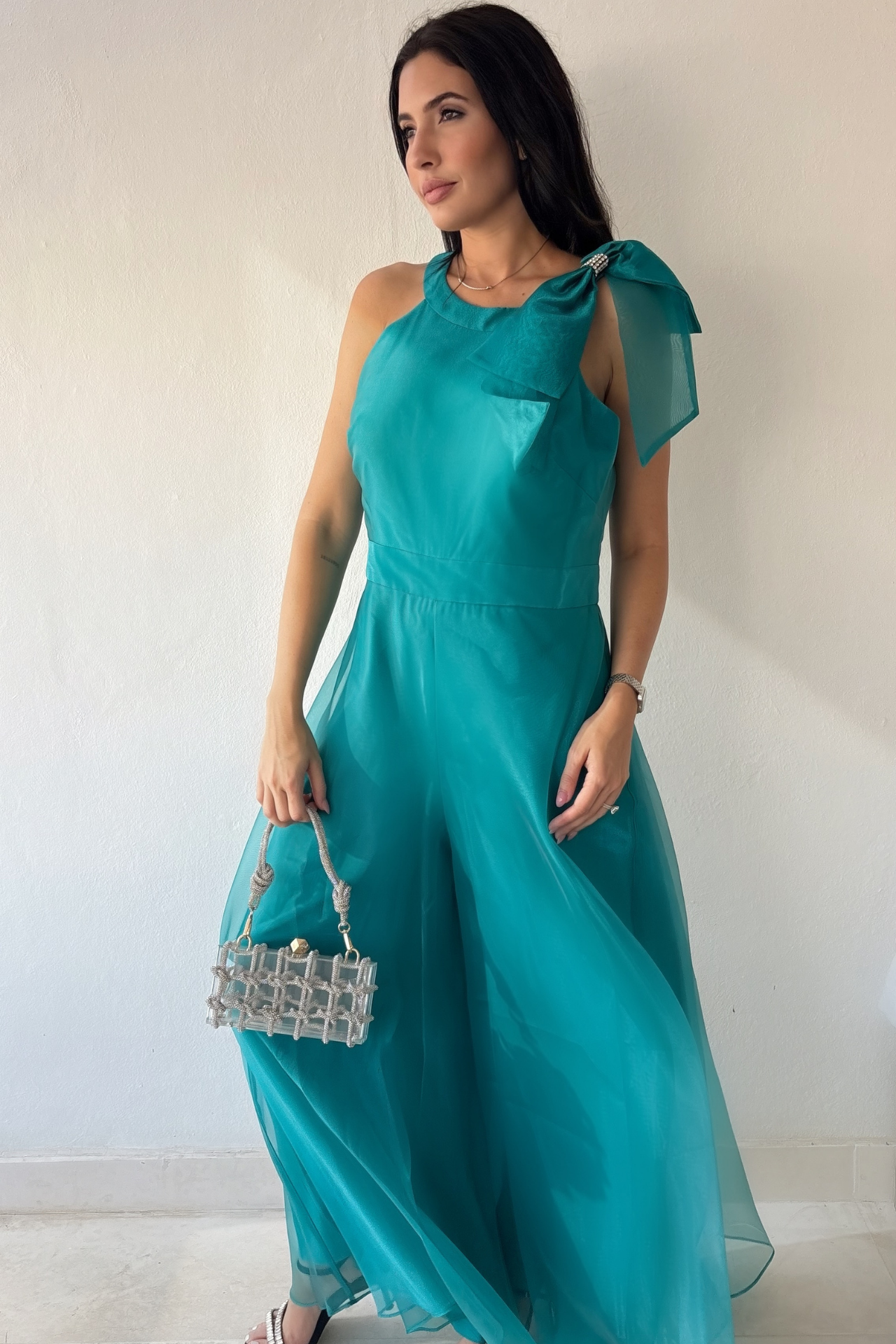 Woman wearing the Aurora Statement Jumpsuit in teal with shoulder bow, wide-leg silhouette and sheer flowing overlay.