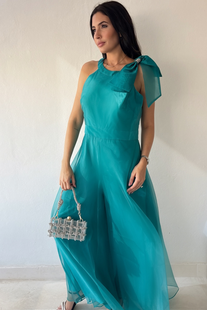 Woman wearing the Aurora Statement Jumpsuit in teal with shoulder bow, wide-leg silhouette and sheer flowing overlay.