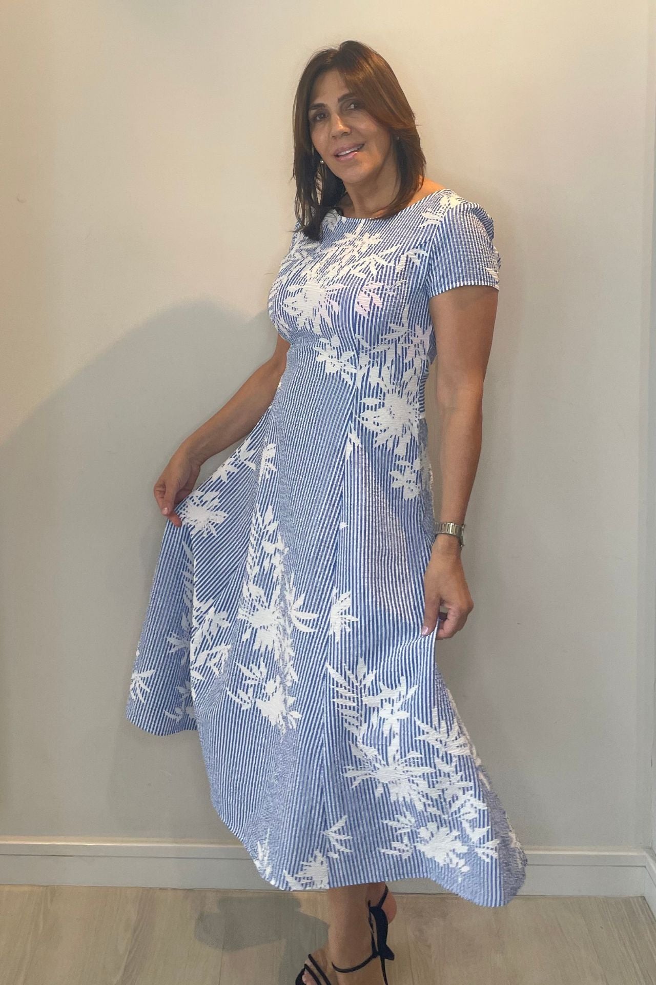 Woman wearing a blue and white floral dress against a plain wall.