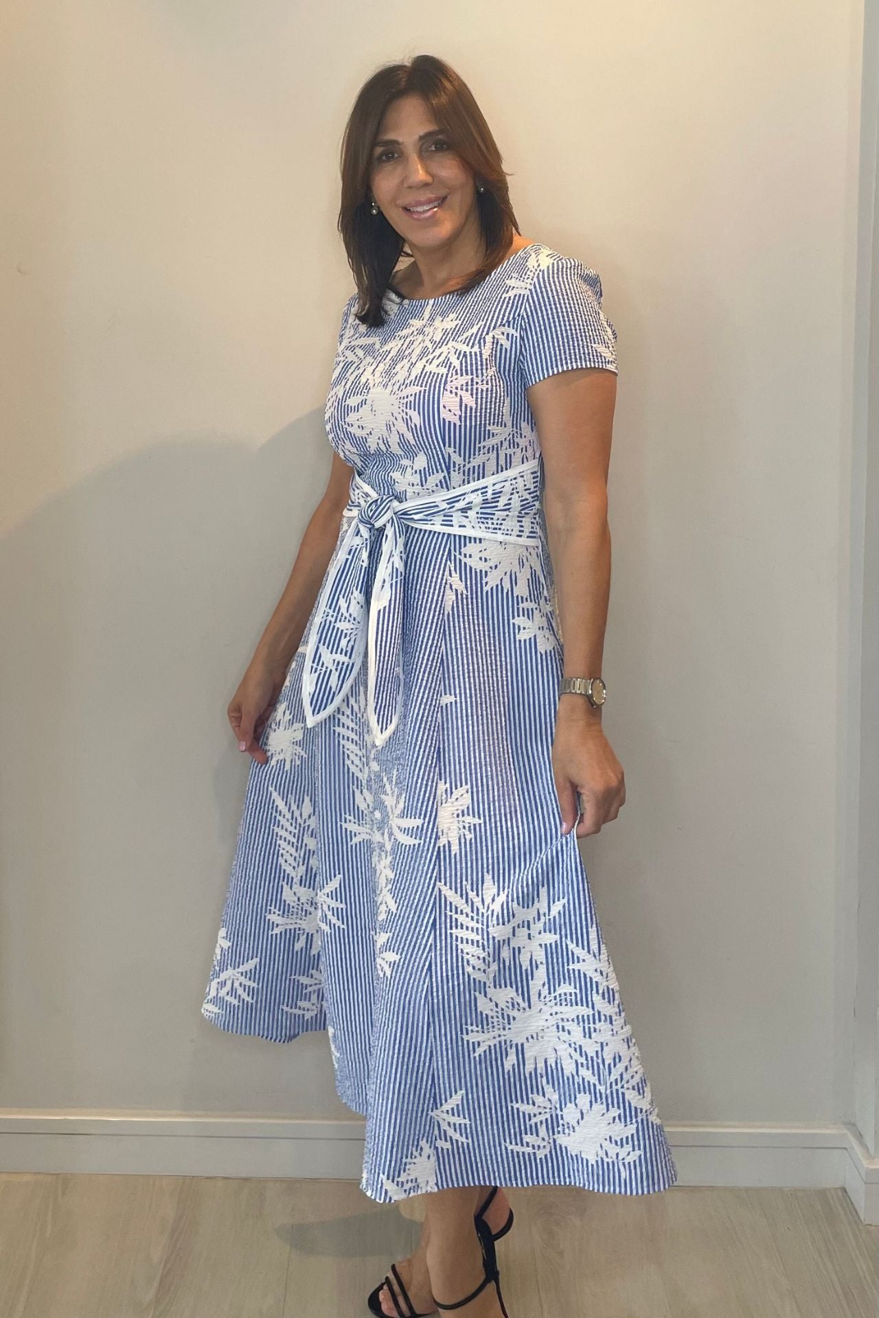 Woman wearing a blue and white floral dress against a plain wall.