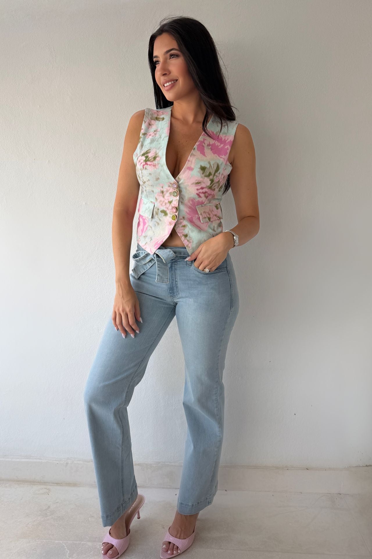 Model wearing the Blush Bloom Vest featuring a pink floral print, V-neckline, and front button closure styled with light-wash denim.