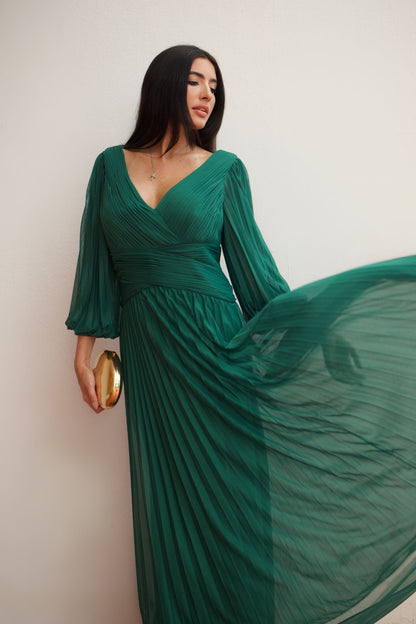 Model wearing an emerald green pleated wrap formal gown with long sleeves and flowing skirt.
