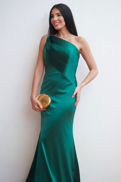 Model wearing an emerald green one-shoulder satin column gown with draped detailing, elegant silhouette, and floor-length design, styled with a gold clutch.