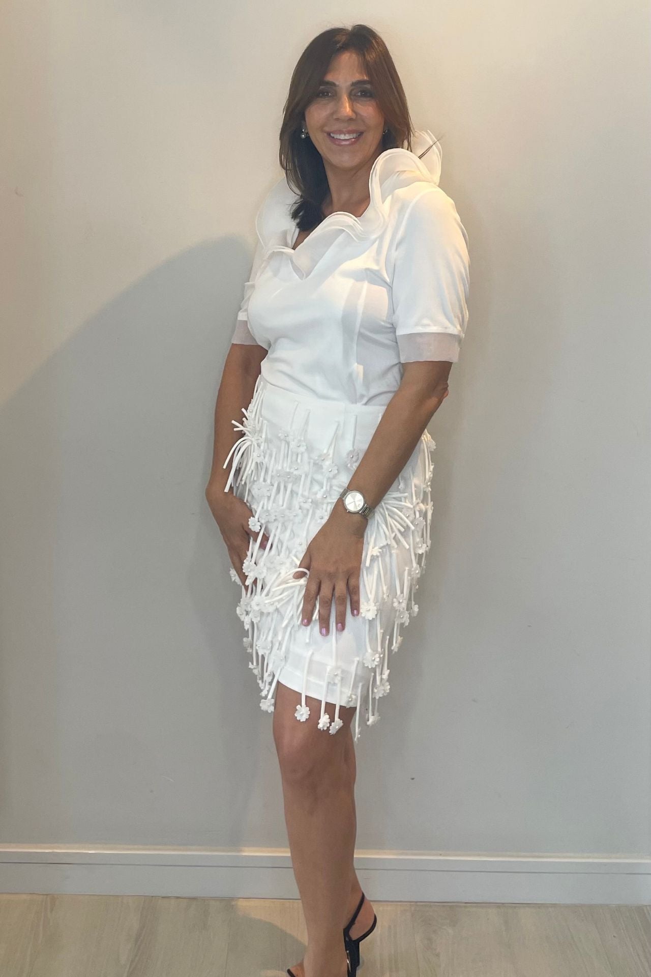 Woman wearing a white set with fringe details against a plain wall.