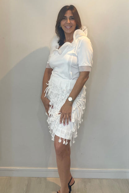 Woman wearing a white set with fringe details against a plain wall.
