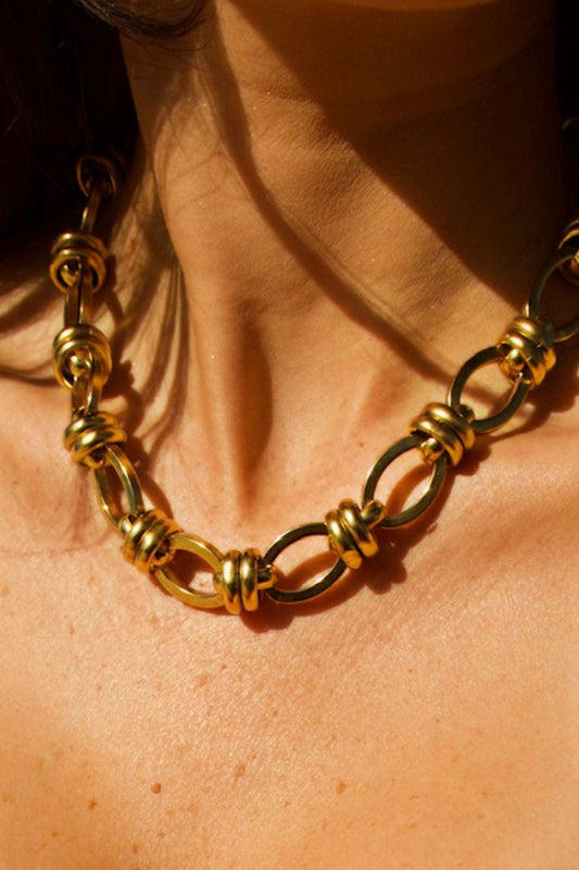 A close-up image of a woman's neck wearing a short gold-tone brass necklace with an oval link design.
