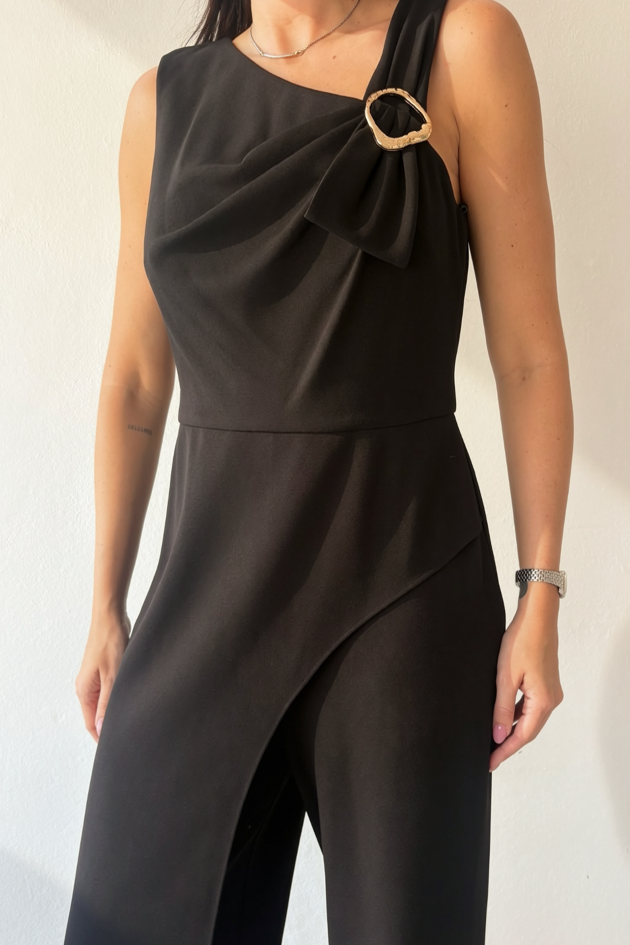 Woman wearing the Midnight Grace Jumpsuit, a black wide-leg jumpsuit with asymmetrical draping, gold-tone shoulder buckle and front overlay.