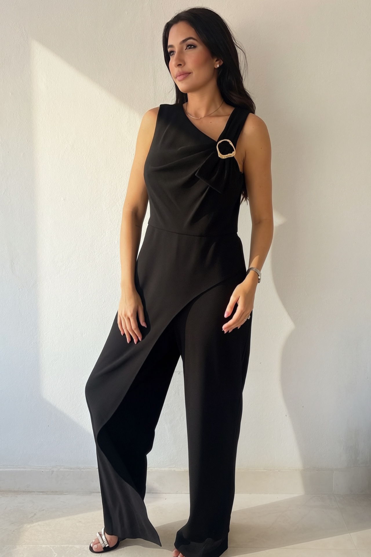 Woman wearing the Midnight Grace Jumpsuit, a black wide-leg jumpsuit with asymmetrical draping, gold-tone shoulder buckle and front overlay.