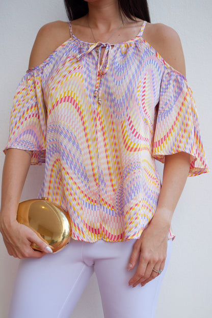 Woman wearing a multicolor mosaic off-shoulder top with cold-shoulder sleeves and a relaxed fit, paired with light-colored pants.