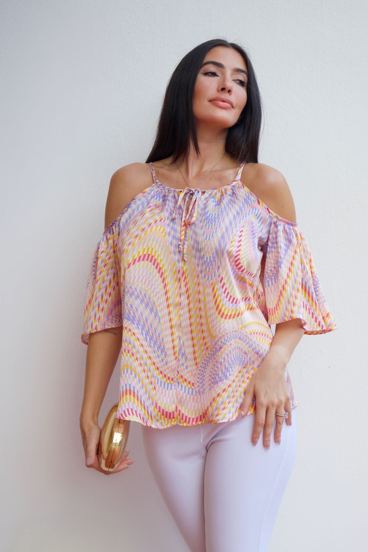 Woman wearing a multicolor mosaic off-shoulder top with cold-shoulder sleeves and a relaxed fit, paired with light-colored pants.