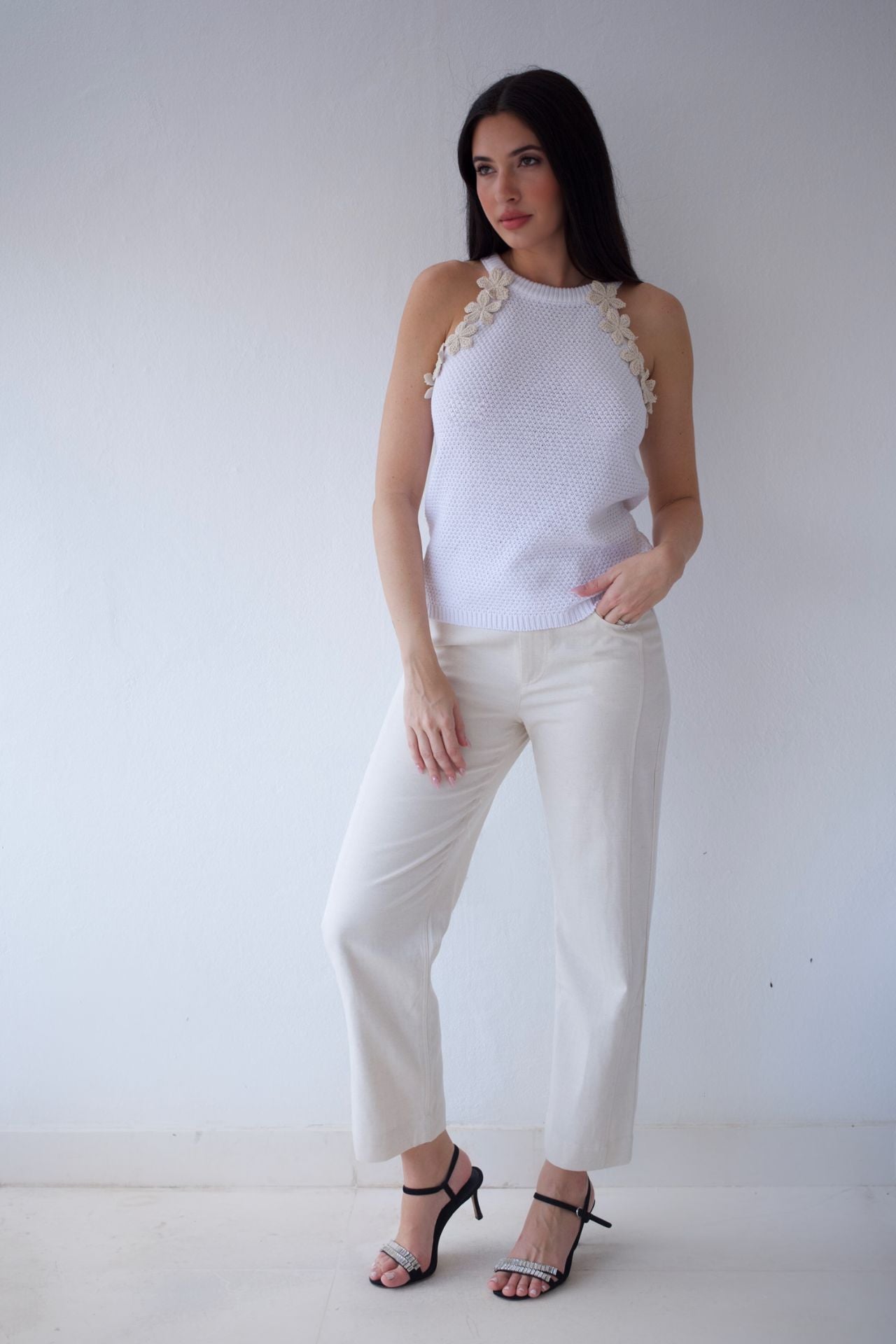 Woman wearing a white sleeveless top with crochet beige flower details and natural pants against a plain background