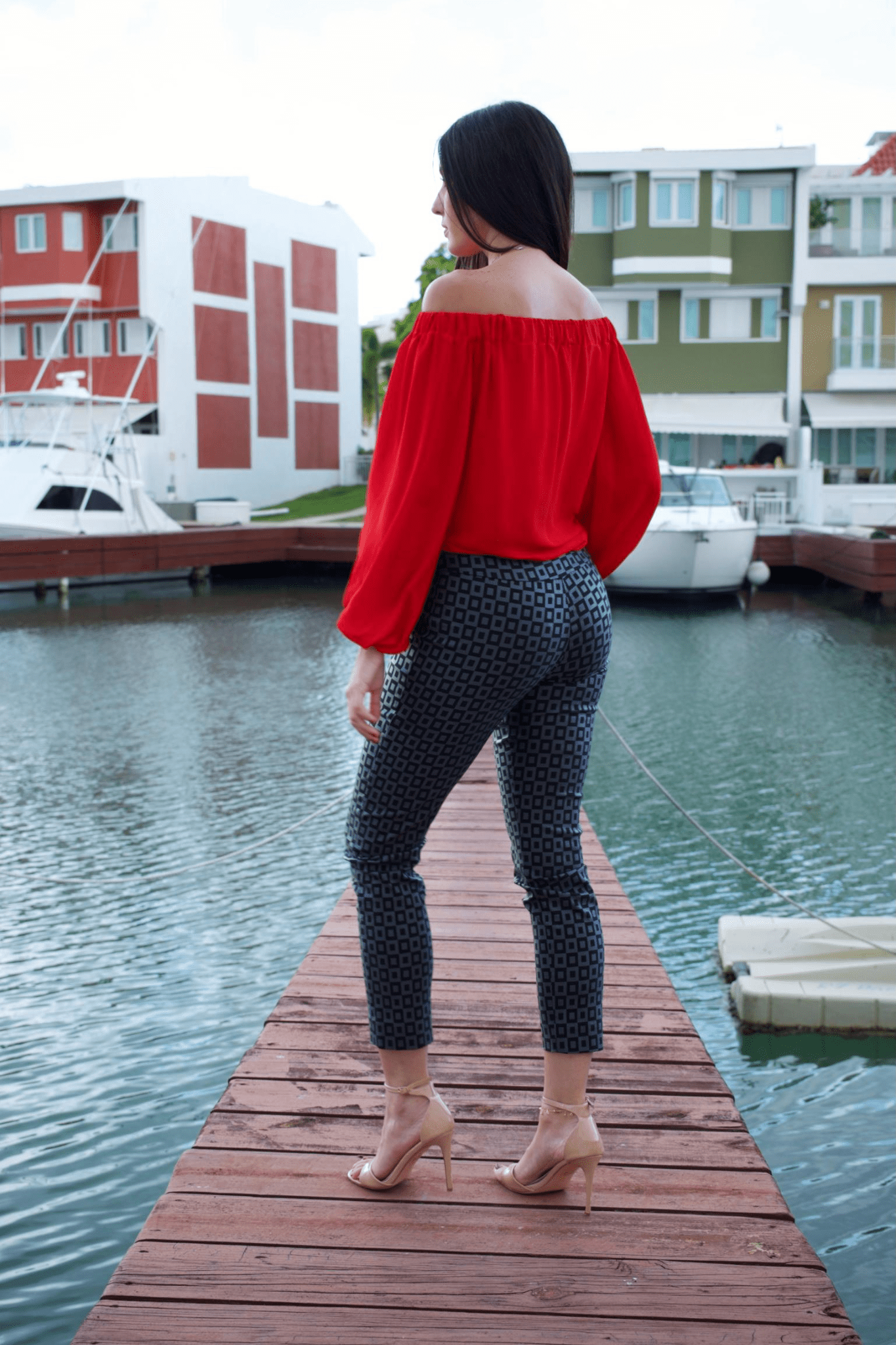 A woman wearing a vibrant red, off-shoulder long sleeve top with a crew neckline, paired with black patterned pants. The top features a loose fit and long bubble sleeves.