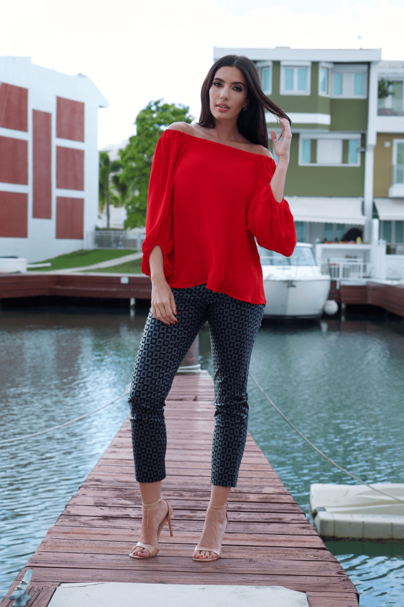 A woman wearing a vibrant red, off-shoulder long sleeve top with a crew neckline, paired with black patterned pants. The top features a loose fit and long bubble sleeves.