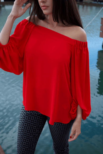 A woman wearing a vibrant red, off-shoulder long sleeve top with a crew neckline, paired with black patterned pants. The top features a loose fit and long bubble sleeves.