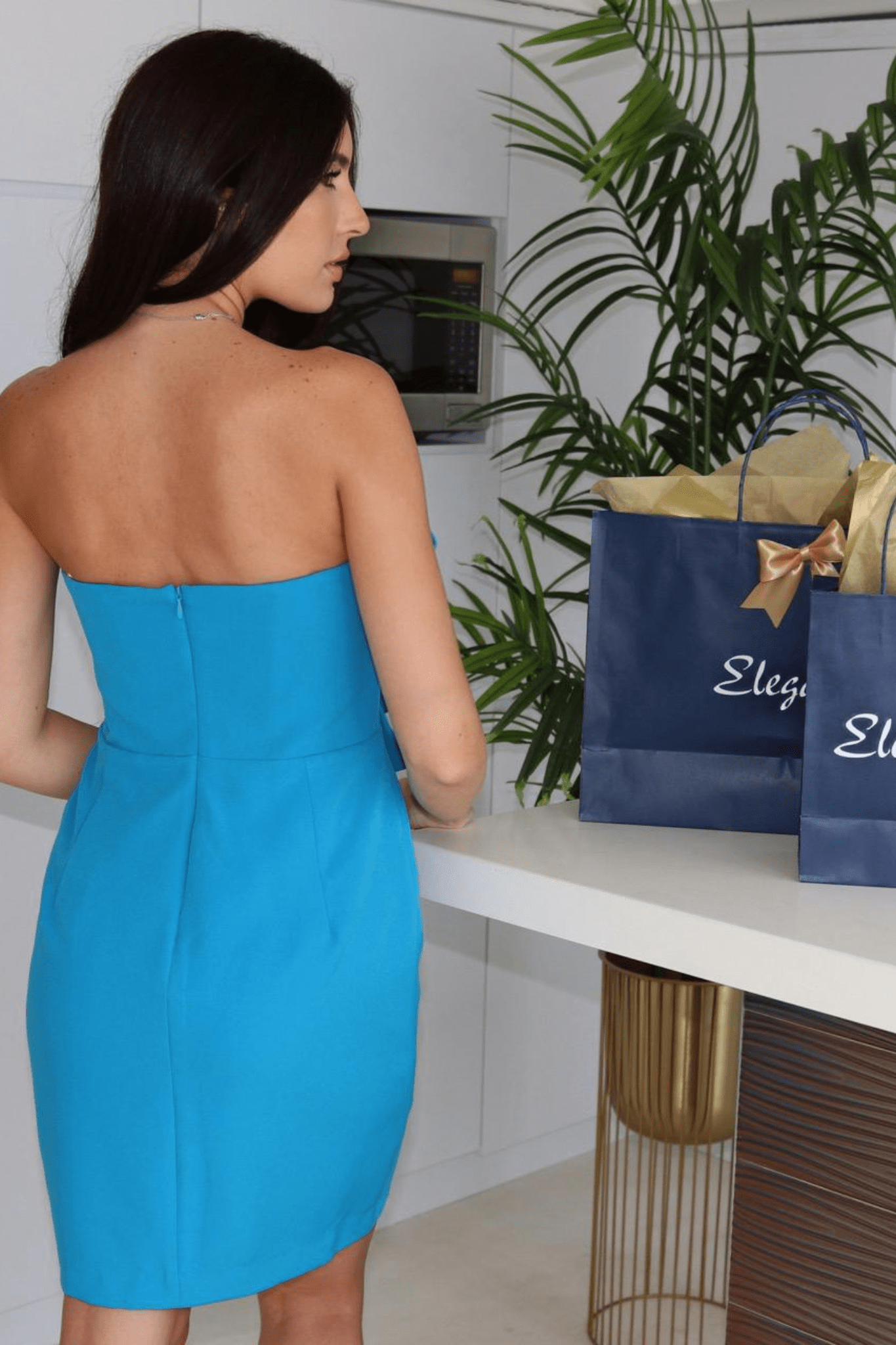 Back view of a woman wearing a strapless, turquoise cocktail dress with a large flower detail at the front.