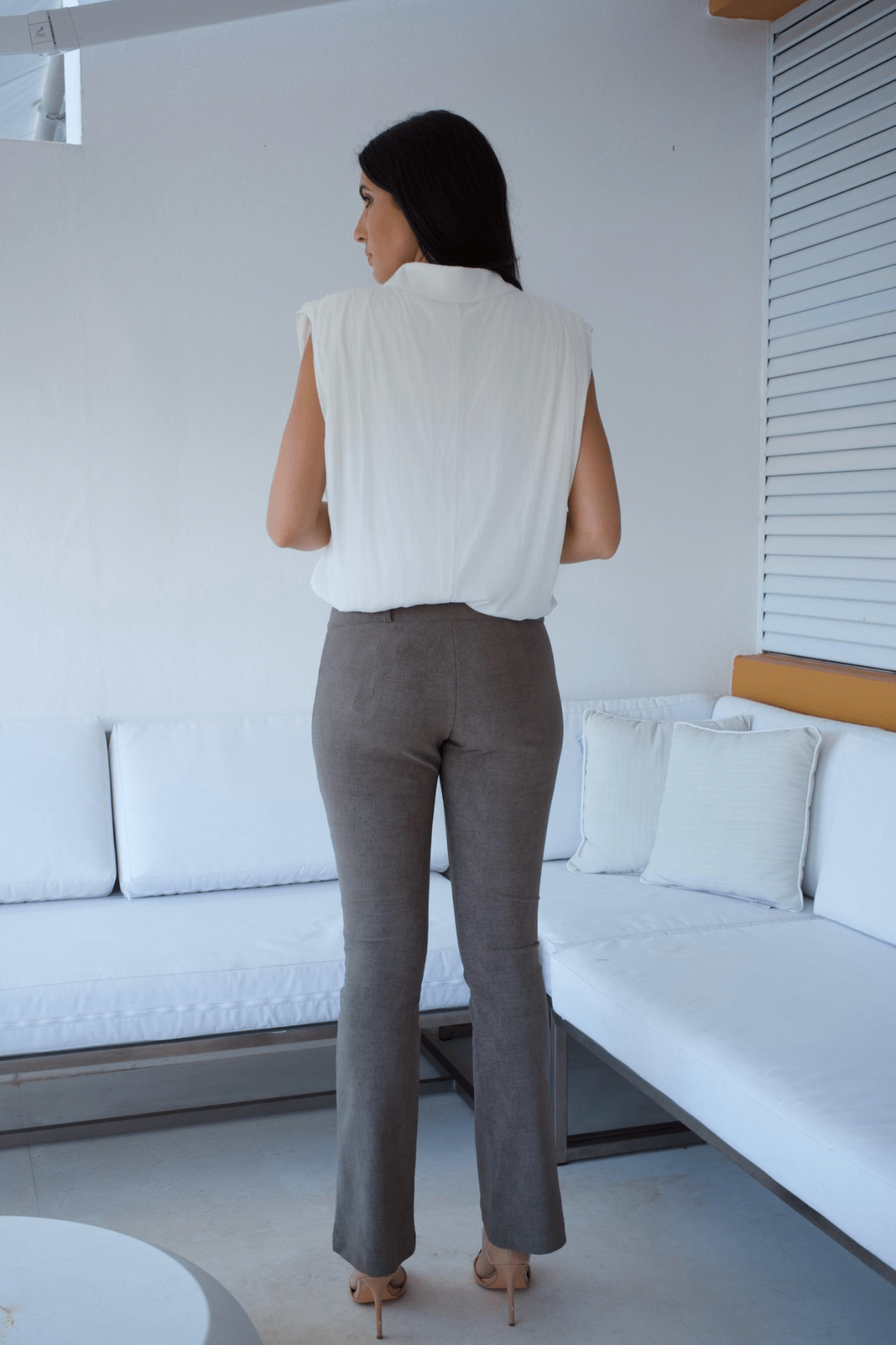 A woman standing in a living room wearing taupe flare pants and a white sleeveless top with a bubble hem.