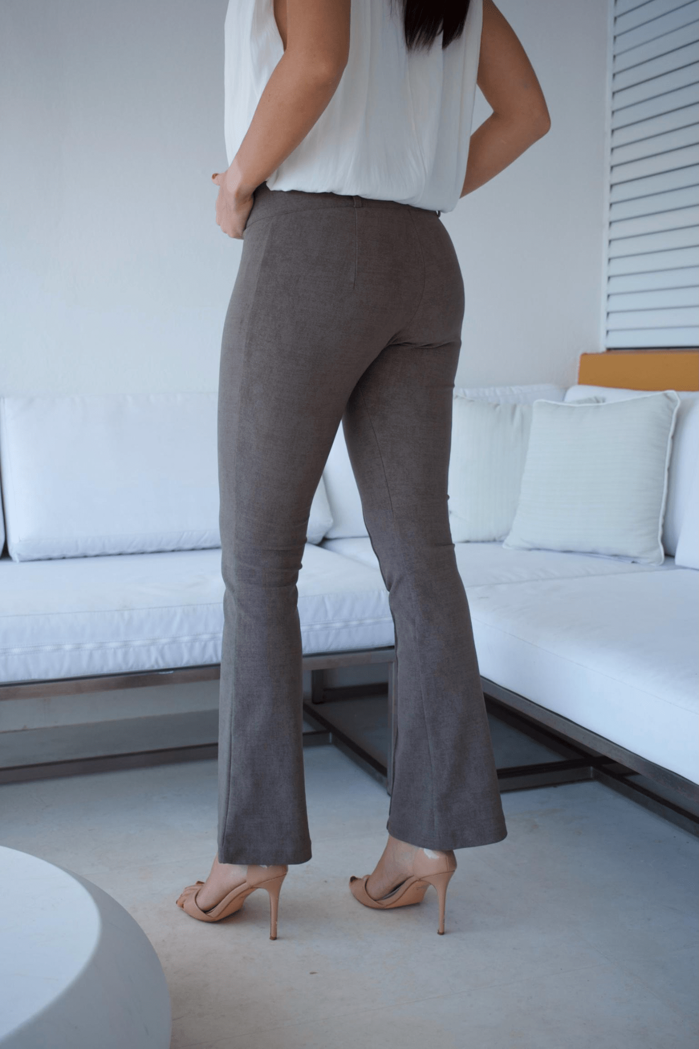 A woman standing in a living room wearing taupe flare pants and a white sleeveless top with a bubble hem.