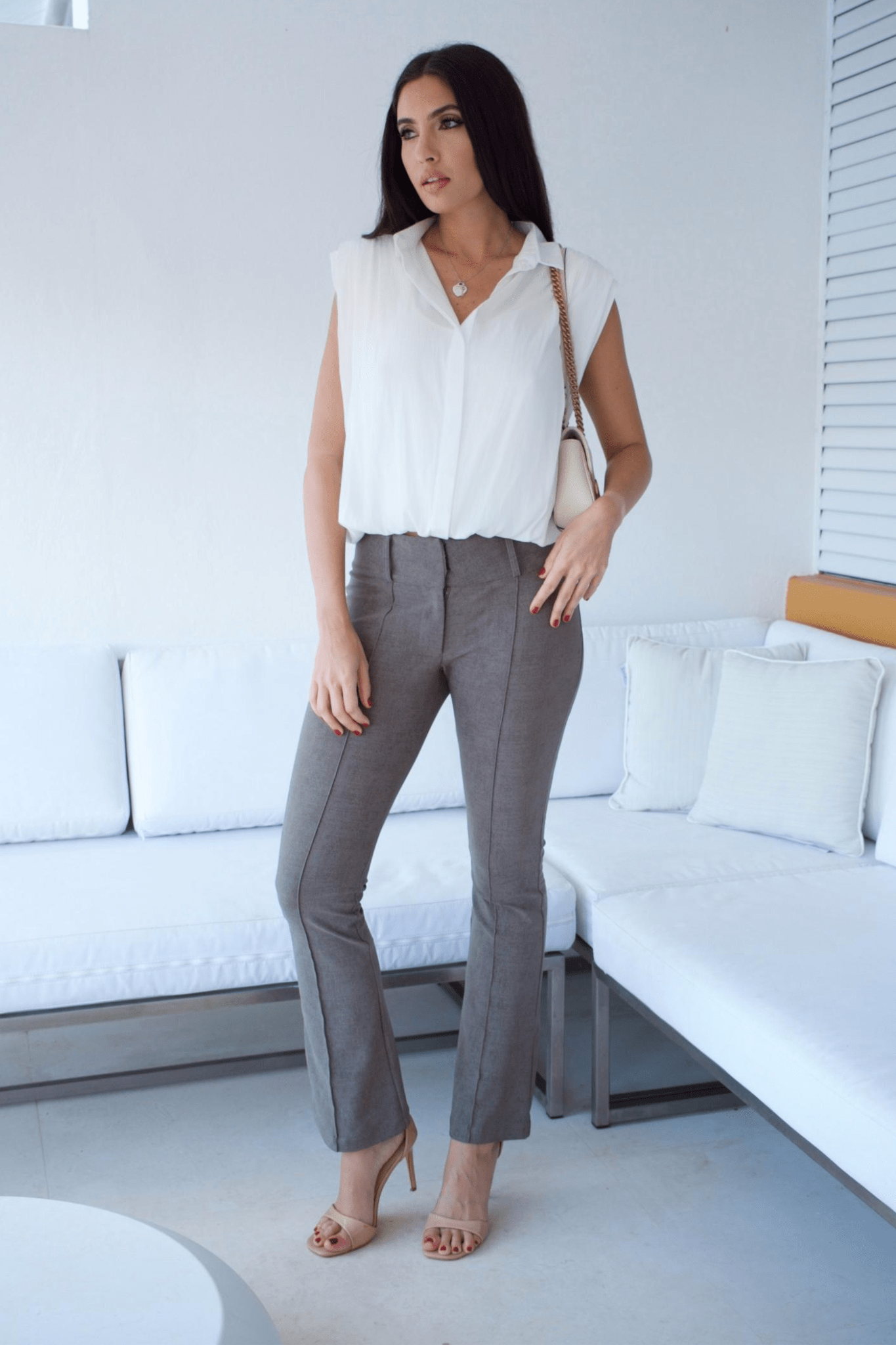 A woman standing in a living room wearing taupe flare pants and a white sleeveless top with a bubble hem.