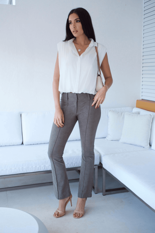 A woman standing in a living room wearing taupe flare pants and a white sleeveless top with a bubble hem.