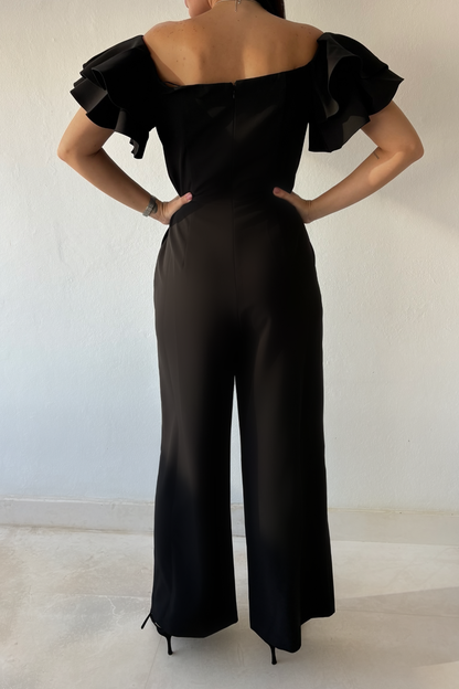 Woman wearing the Timeless Noir Jumpsuit, a black wide-leg jumpsuit with square neckline, fitted waist and statement layered ruffle sleeves.