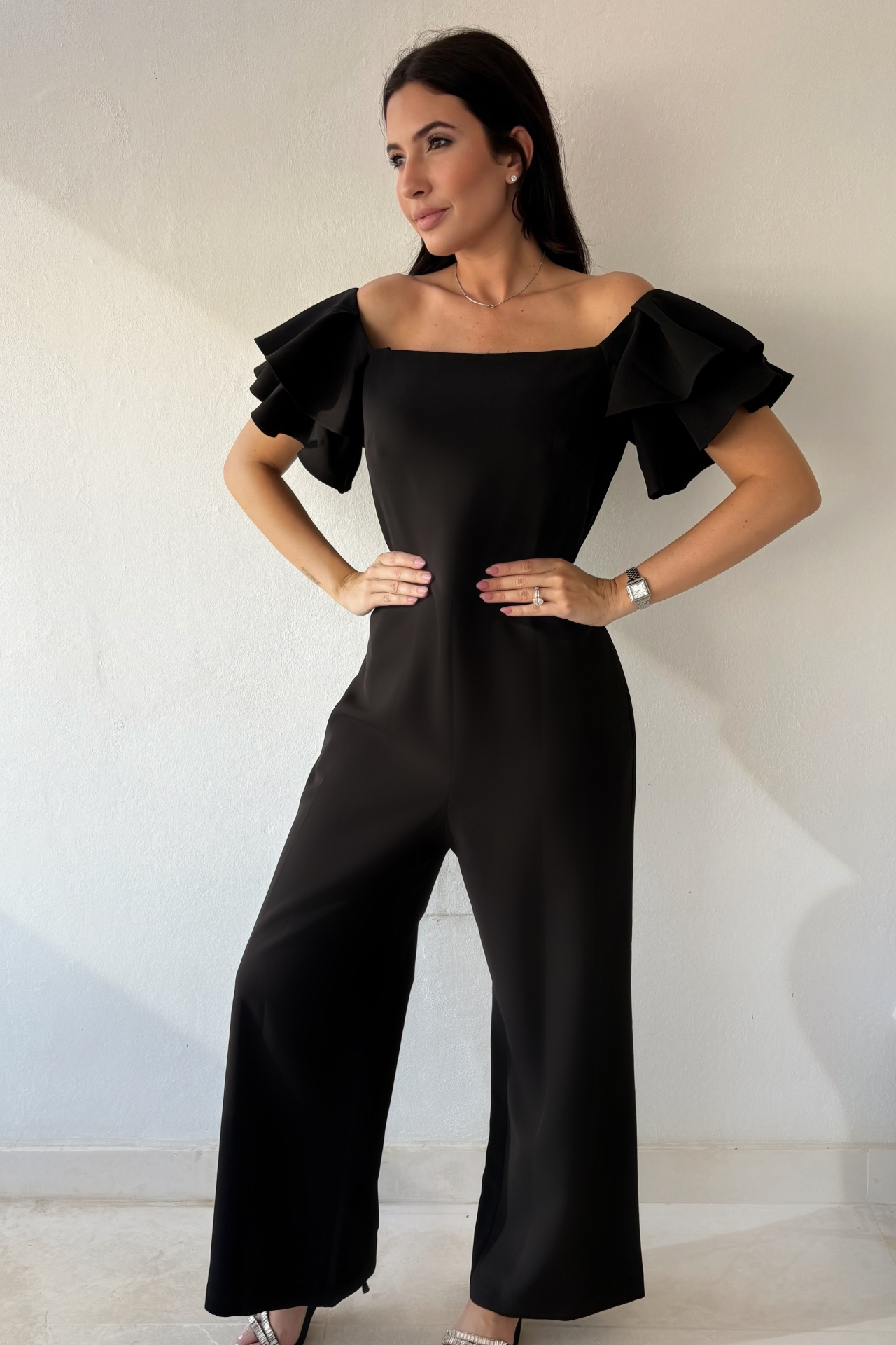 Woman wearing the Timeless Noir Jumpsuit, a black wide-leg jumpsuit with square neckline, fitted waist and statement layered ruffle sleeves.