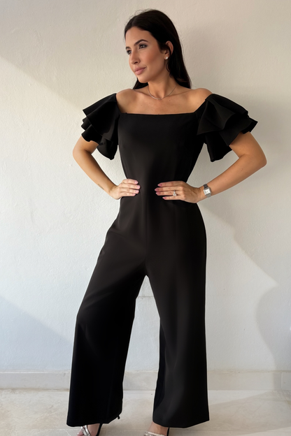 Woman wearing the Timeless Noir Jumpsuit, a black wide-leg jumpsuit with square neckline, fitted waist and statement layered ruffle sleeves.