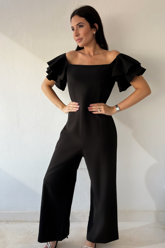 Woman wearing the Timeless Noir Jumpsuit, a black wide-leg jumpsuit with square neckline, fitted waist and statement layered ruffle sleeves.