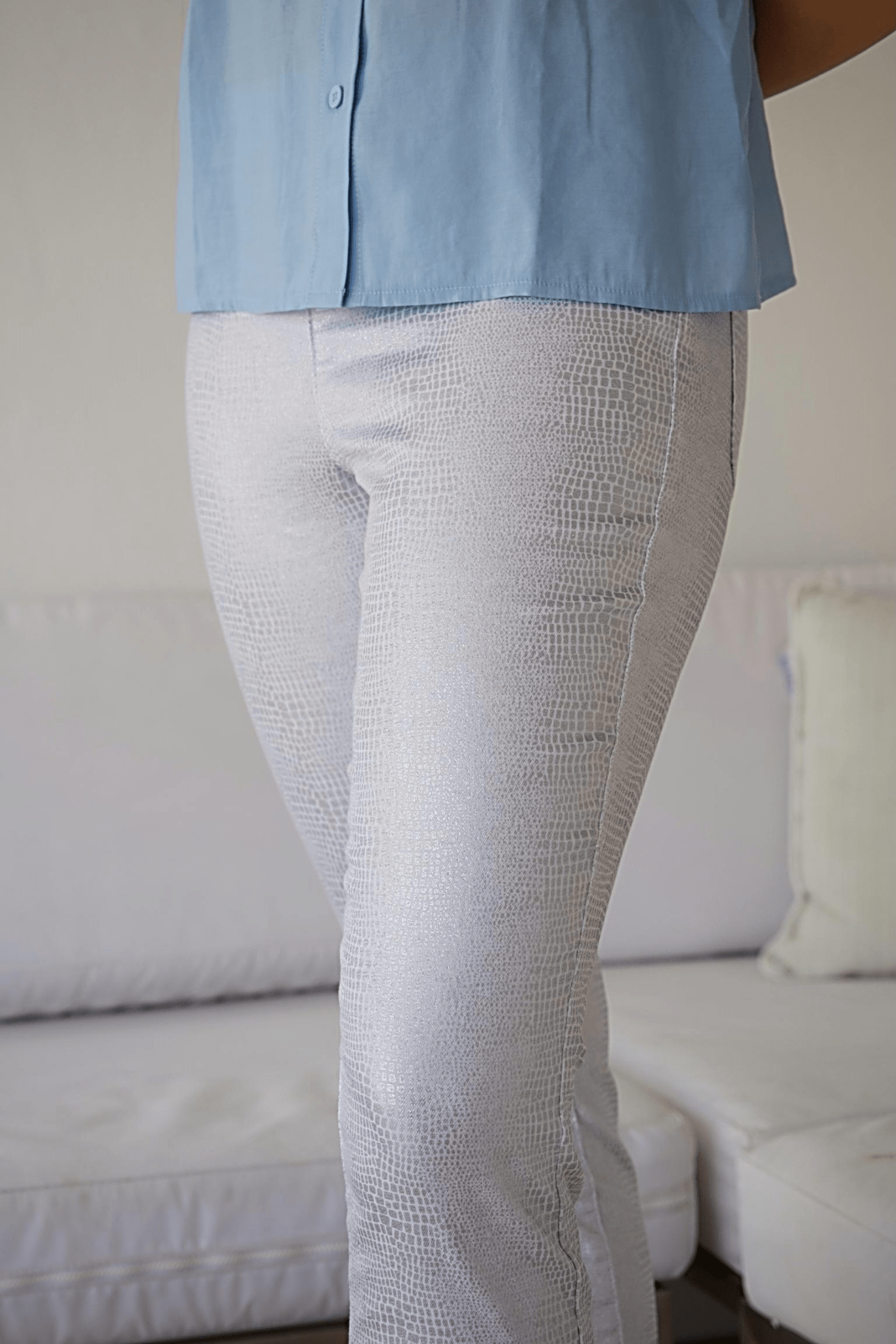 A woman standing in a modern living room wearing white crop pants with a silver snake print and a blue top with ruffle sleeves.