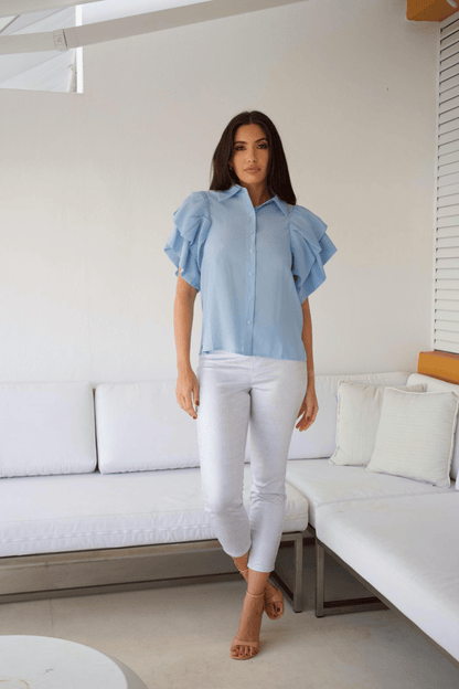 A woman standing in a modern living room wearing white crop pants with a silver snake print and a blue top with ruffle sleeves. 