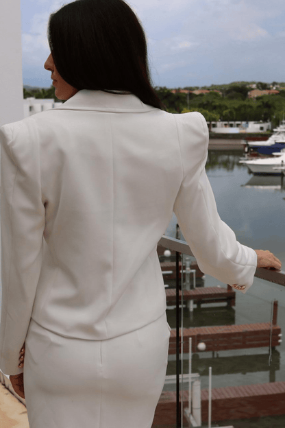  woman wearing a white short skirt set with gold buttons, including a long sleeve jacket and a skirt, standing by a railing with a boatyard in the background.