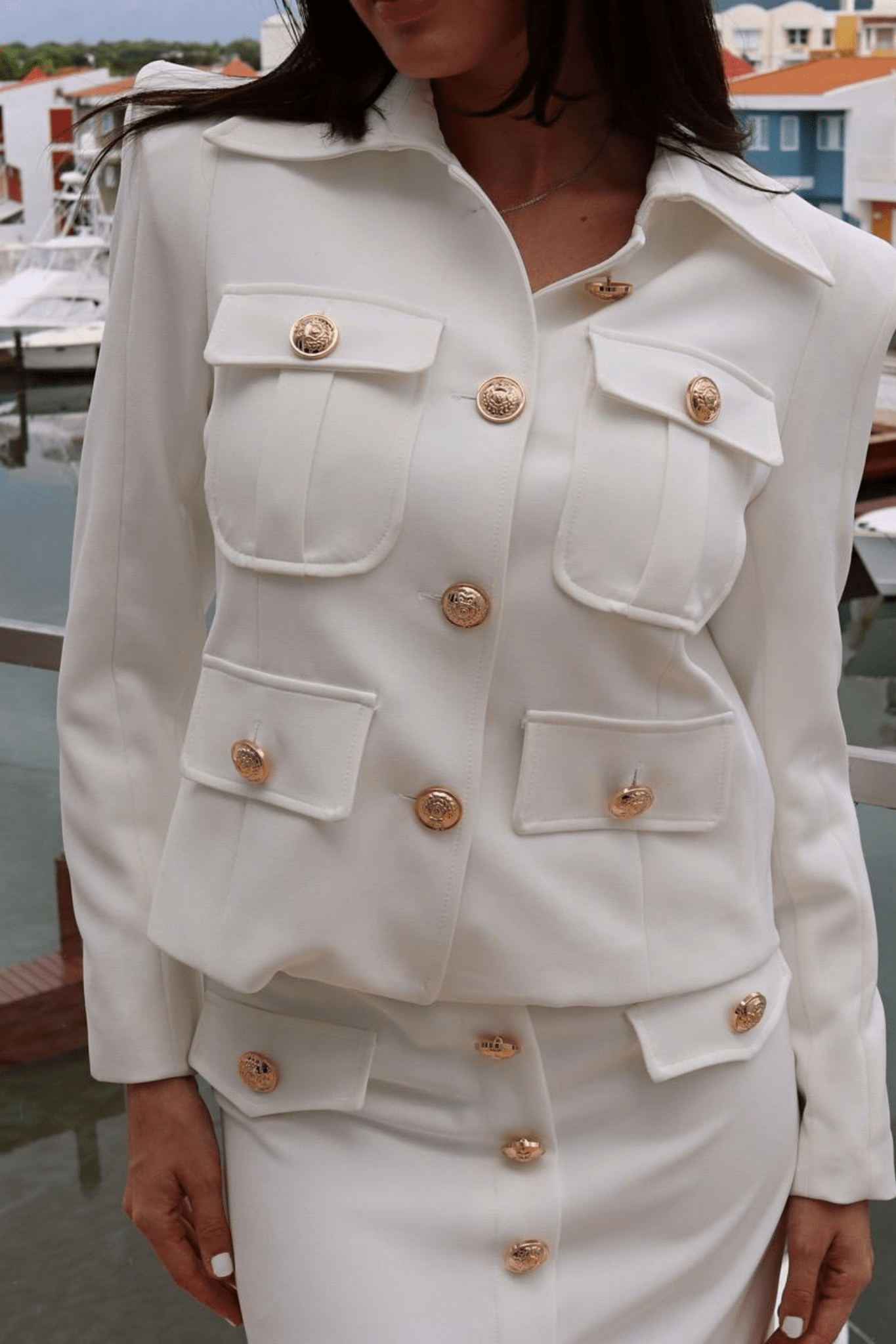 woman wearing a white short skirt set with gold buttons, including a long sleeve jacket and a skirt, standing by a railing with a boatyard in the background.