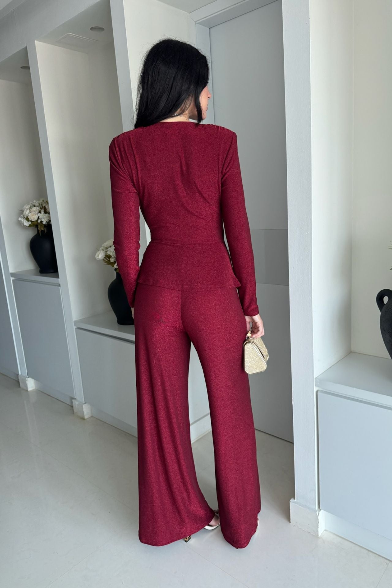 Back view of a woman wearing a burgundy long-sleeve knot-detail wide-leg jumpsuit standing indoors.