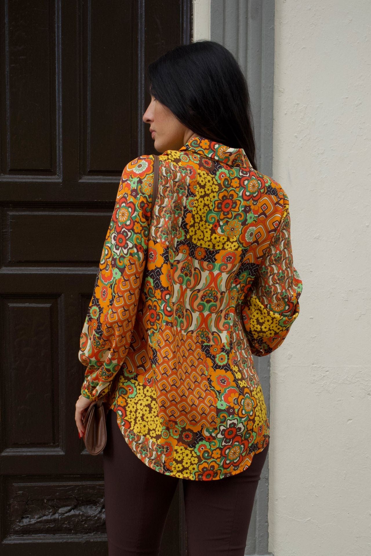 Back view of a woman wearing retro patchwork print blouse with long sleeves and chocolate brown slim pants.