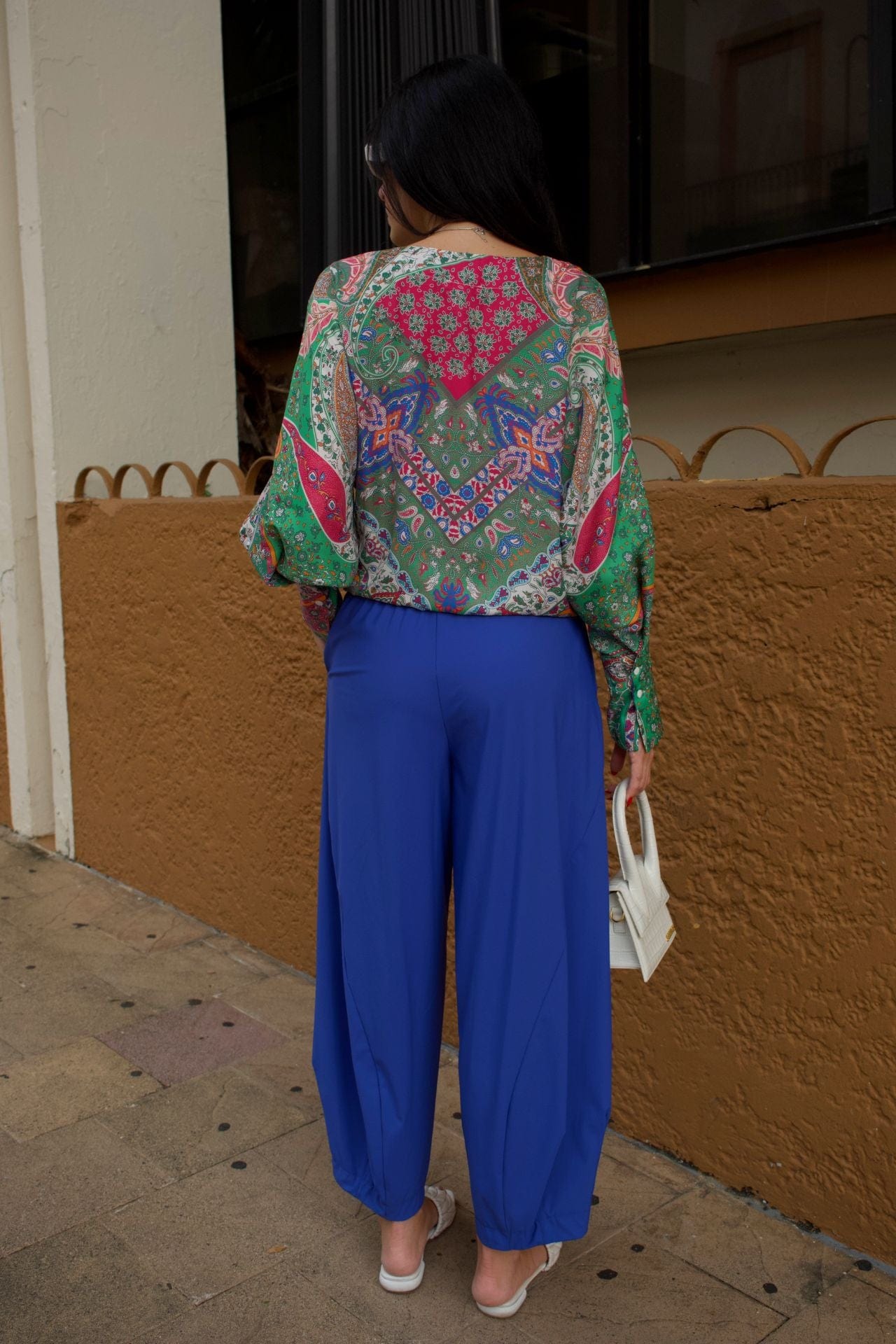 Back view of a colorful printed V-neck shirt with balloon sleeves and front tie hem styled with cobalt blue pants and white sandals