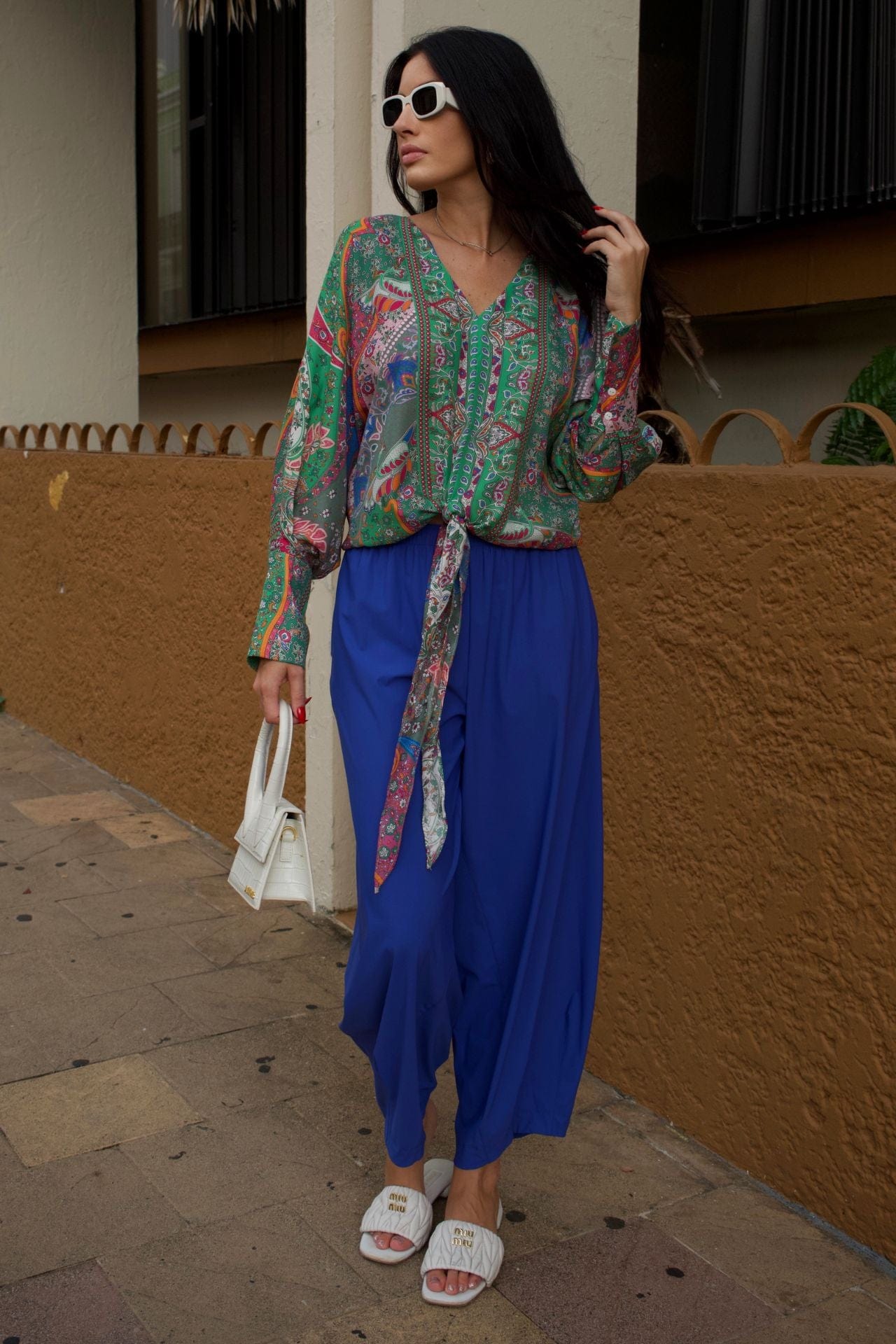 Model wearing colorful printed V-neck shirt with balloon sleeves and front tie hem styled with cobalt blue pants and white sandals.